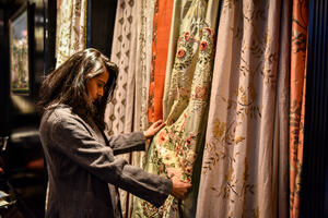 A woman in a gray jacket holds a gold floral fabric sample hanging amongst various colorful, patterned fabrics.